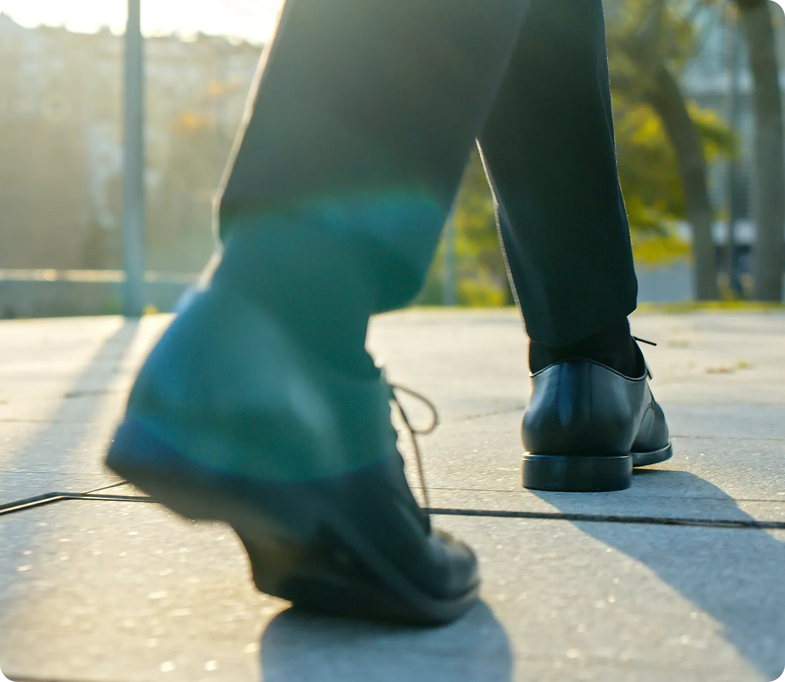 Close-up of a person walking on a sunny day, in black dress shoes and dark pants. The low angle and lighting create a dynamic, forward-moving feel.